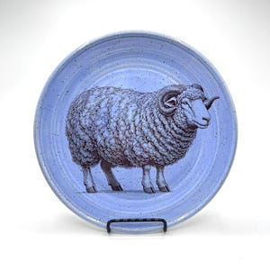 May include: A round, light blue ceramic plate with a detailed illustration of a ram. The ram is depicted in shades of blue and gray, with a textured, woolly appearance. The plate has a speckled finish and is displayed on a black metal stand.