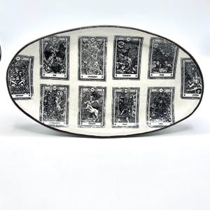 May include: White oval ceramic serving dish with a black border. The dish is decorated with black and white images of tarot cards. The cards are arranged in a grid pattern and include the Empress, Judgement, Strength, Star, Lovers, Chariot, Magician, Moon, Fool, and World.