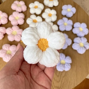 May include: A collection of handmade, plush flower decorations in various colors. The flowers have white, pink, and lavender petals with yellow or orange centers. The flowers are arranged on a wooden surface.