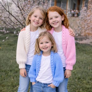 May include: Three young girls wearing white t-shirts and knitted cardigans in cream, pink, and blue. The cardigans have a cable knit design. They are smiling and standing outside in front of a flowering tree.