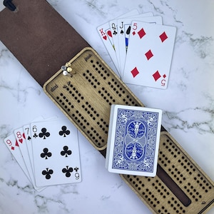 Può includere: Un tabellone di cribbage in legno con una custodia di trasporto in pelle marrone. Il tabellone ha un'etichetta "start" e "finish". Il tabellone è aperto e mostra i pioli e i buchi. Ci sono carte da gioco sul tabellone e accanto ad esso. Le carte includono un 5 di quadri, un jack di picche, una regina di cuori, un re di fiori e un jolly.