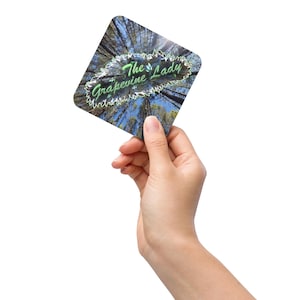 May include: A square coaster with rounded corners, held in a hand. The coaster features a forest scene with the text "The Grapevine Lady" in green script. The background shows tall trees and a blue sky.
