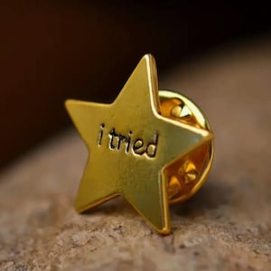Puede incluir: Un pin de estrella dorada con las palabras "i tried" inscritas en él.