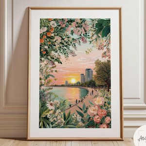 May include: A framed art print depicting a sunset over a coastal city skyline, framed by lush green foliage and pink flowers. The scene features a curved shoreline, a paved walkway with small figures, and tall buildings under a soft orange sky.