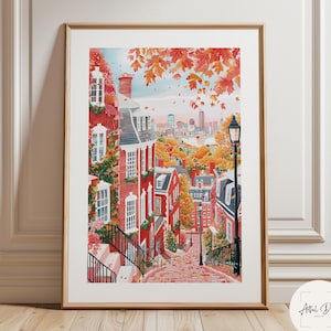 May include: Framed watercolor print of a charming street scene in autumn. Red brick buildings with white trim line a cobblestone street, with fall foliage in shades of orange and red. A cityscape is visible in the distance. The print is in a gold frame.