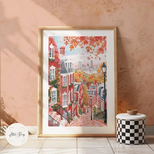 May include: Framed watercolor print of a charming red brick street scene with autumn foliage. The artwork depicts a sloping street lined with buildings, a lamppost, and a distant cityscape. The print is in a gold frame and leans against a peach-colored wall.