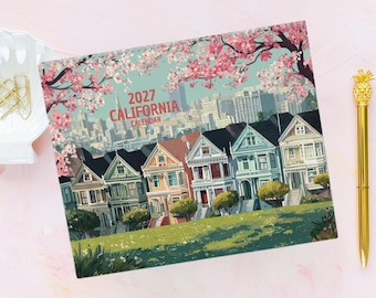 May include: A 2027 California calendar featuring a colorful illustration of Victorian houses in San Francisco under a canopy of pink cherry blossoms. The scene includes a city skyline in the background and a green grassy foreground with a gold pen nearby.