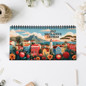Op de afbeelding: Een spiraalgebonden 2027 Zuid-Afrika kalender met een kleurrijke illustratie van een kuststad met levendige huizen, een vuurtoren, bergen en inheemse proteabloemen op de voorgrond onder een zachte blauwe lucht met wolken.
