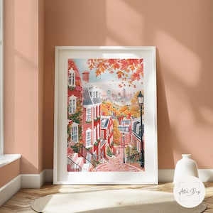 May include: A framed watercolor print of a charming cityscape in autumn. Red brick buildings with white trim line a cobblestone street, with fall foliage in shades of orange and red. A distant skyline is visible under a blue sky.