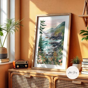 May include: A framed watercolor print of a coastal landscape with a wooden frame. The artwork depicts a rocky shoreline, turquoise water, and lush green hills. The print is displayed on a wooden cabinet with a vintage radio and a potted plant.