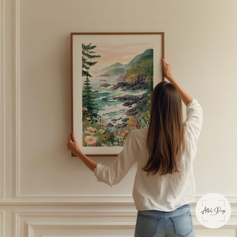 May include: A framed watercolor print of a coastal landscape with a woman holding it up. The artwork depicts a rocky shoreline, ocean waves, and a forest. The color palette includes greens, blues, and soft pinks. The frame is a warm brown.