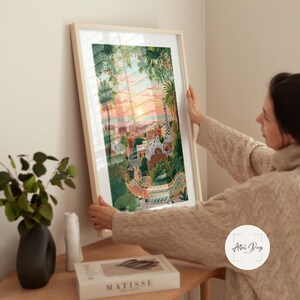 Pode incluir: Uma impress&atilde;o de arte emoldurada com uma ilustra&ccedil;&atilde;o colorida de uma paisagem urbana ao p&ocirc;r do sol. A obra de arte apresenta edif&iacute;cios, &aacute;rvores e uma ponte, com uma paleta de cores quentes de laranjas, rosas e verdes. A impress&atilde;o &eacute; segurada por uma pessoa.