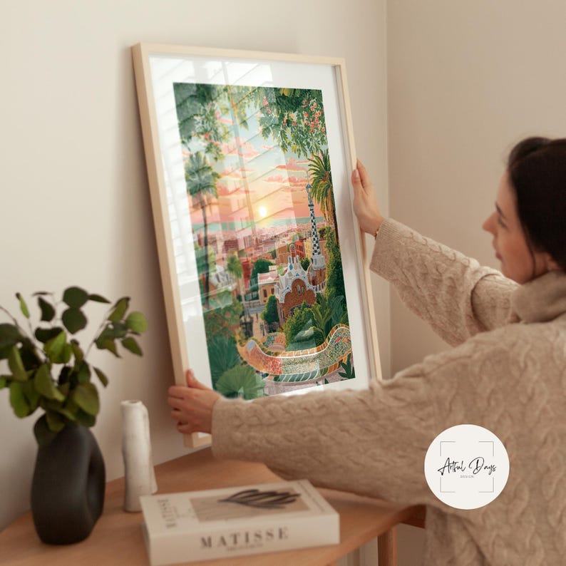 Pode incluir: Uma impress&atilde;o de arte emoldurada com uma ilustra&ccedil;&atilde;o colorida de uma paisagem urbana ao p&ocirc;r do sol. A obra de arte apresenta edif&iacute;cios, &aacute;rvores e uma ponte, com uma paleta de cores quentes de laranjas, rosas e verdes. A impress&atilde;o &eacute; segurada por uma pessoa.