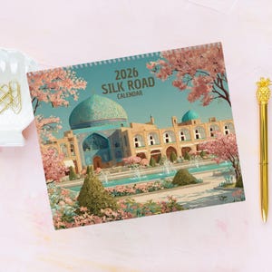 May include: A 2026 Silk Road calendar featuring a detailed illustration of a Middle Eastern architectural landmark with a turquoise dome and fountains. Pink flowering trees frame the scene, with a gold pen in the background.