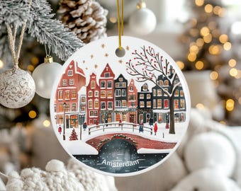 Peut inclure: Une boule de Noël ronde en céramique avec une illustration aquarelle d'un paysage urbain d'Amsterdam enneigé avec des bâtiments colorés, un pont et le texte "Amsterdam".