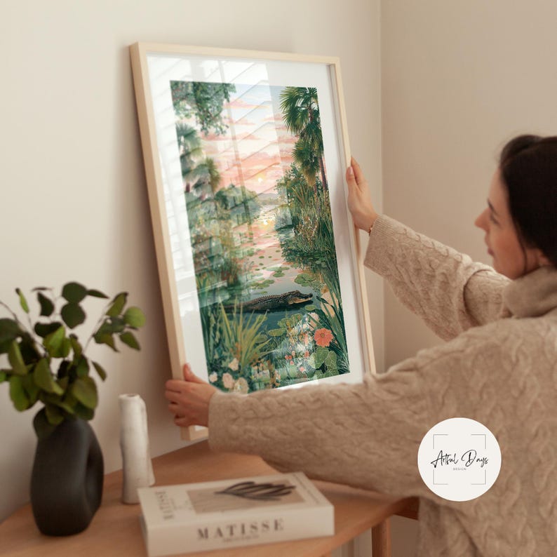 Puede incluir: Una impresi&oacute;n art&iacute;stica enmarcada que presenta un paisaje sereno con un cuerpo de agua, vegetaci&oacute;n exuberante y un cielo al atardecer. La obra de arte incluye un caim&aacute;n en el agua. El marco es de color claro.