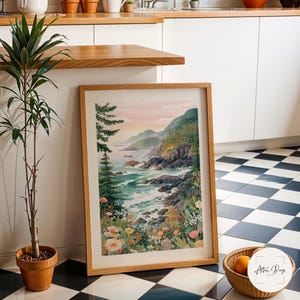 May include: A framed watercolor painting of a coastal landscape with a wooden frame. The artwork depicts a shoreline with crashing waves, lush greenery, and distant mountains under a pastel sky. The painting is leaning against a white wall.