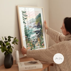 May include: A framed watercolor print of a coastal landscape. The artwork depicts a rocky shoreline with crashing waves, lush greenery, and colorful wildflowers. The frame is a light wood color, and the overall aesthetic is serene and natural.