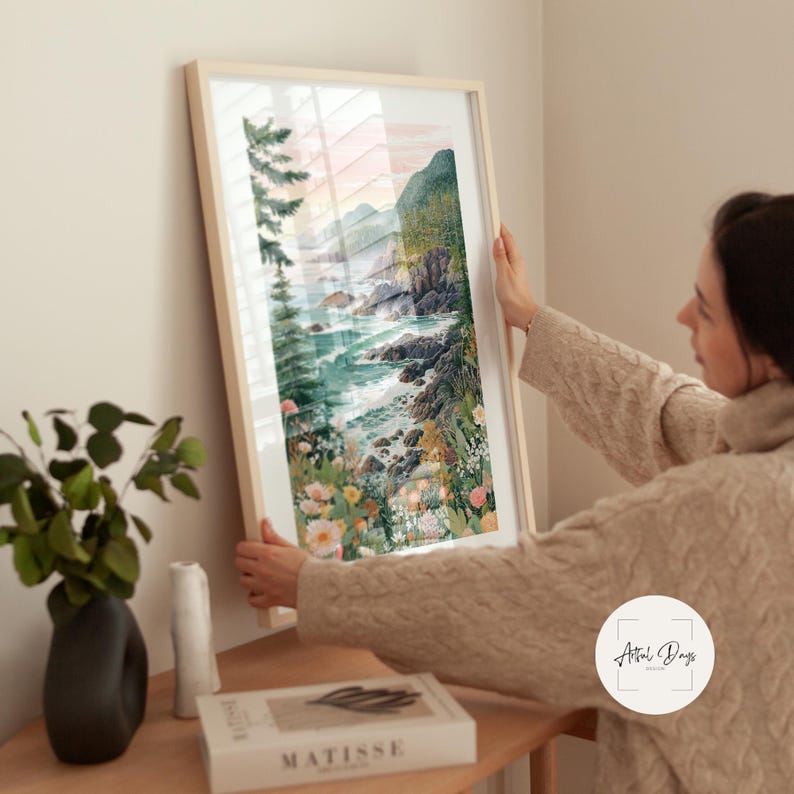 May include: A framed watercolor print of a coastal landscape. The artwork depicts a rocky shoreline with crashing waves, lush greenery, and colorful wildflowers. The frame is a light wood color, and the overall aesthetic is serene and natural.