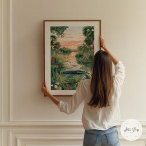 Puede incluir: Una impresi&oacute;n art&iacute;stica enmarcada que representa una serena escena de pantano con una puesta de sol. La obra de arte presenta una masa de agua con un caim&aacute;n, rodeada de exuberante vegetaci&oacute;n y palmeras. El marco es de color marr&oacute;n c&aacute;lido.