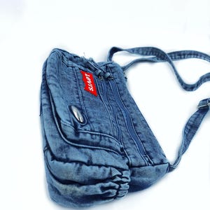 May include: A blue denim crossbody bag featuring a red Levi's brand tag on the front pocket. The bag includes multiple zippered compartments, a silver-toned metal accent, and an adjustable denim shoulder strap with a metal buckle hardware.