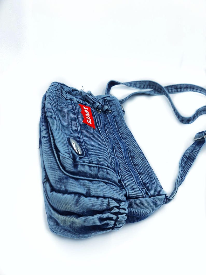 May include: A blue denim crossbody bag featuring a red Levi's brand tag on the front pocket. The bag includes multiple zippered compartments, a silver-toned metal accent, and an adjustable denim shoulder strap with a metal buckle hardware.