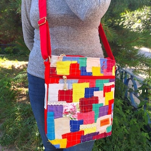 May include: A colorful patchwork crossbody bag with a red strap and zipper. The bag features a variety of fabric squares in red, yellow, blue, and green, with a floral accent and lace trim. The bag is worn over the shoulder.