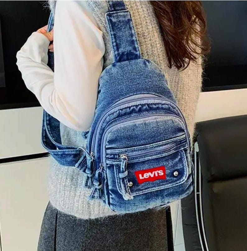 K&ouml;nnte beinhalten: Blaue Jeans-Umh&auml;ngetasche mit mehreren Rei&szlig;verschlussf&auml;chern und einem roten Levi's-Logo-Patch. Die Tasche hat einen einzelnen Schultergurt und eine kleine Tasche auf der Vorderseite. Der Jeansstoff hat einen verblichenen Look.