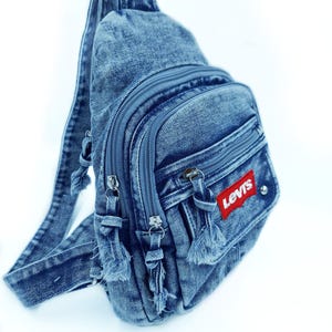 K&ouml;nnte beinhalten: Eine blaue Jeans-Umh&auml;ngetasche mit mehreren Rei&szlig;verschlussf&auml;chern und einem Schultergurt. Die Tasche hat einen roten "Levis"-Aufn&auml;her und dekorative Quasten an den Rei&szlig;verschl&uuml;ssen. Aus Denim gefertigt.