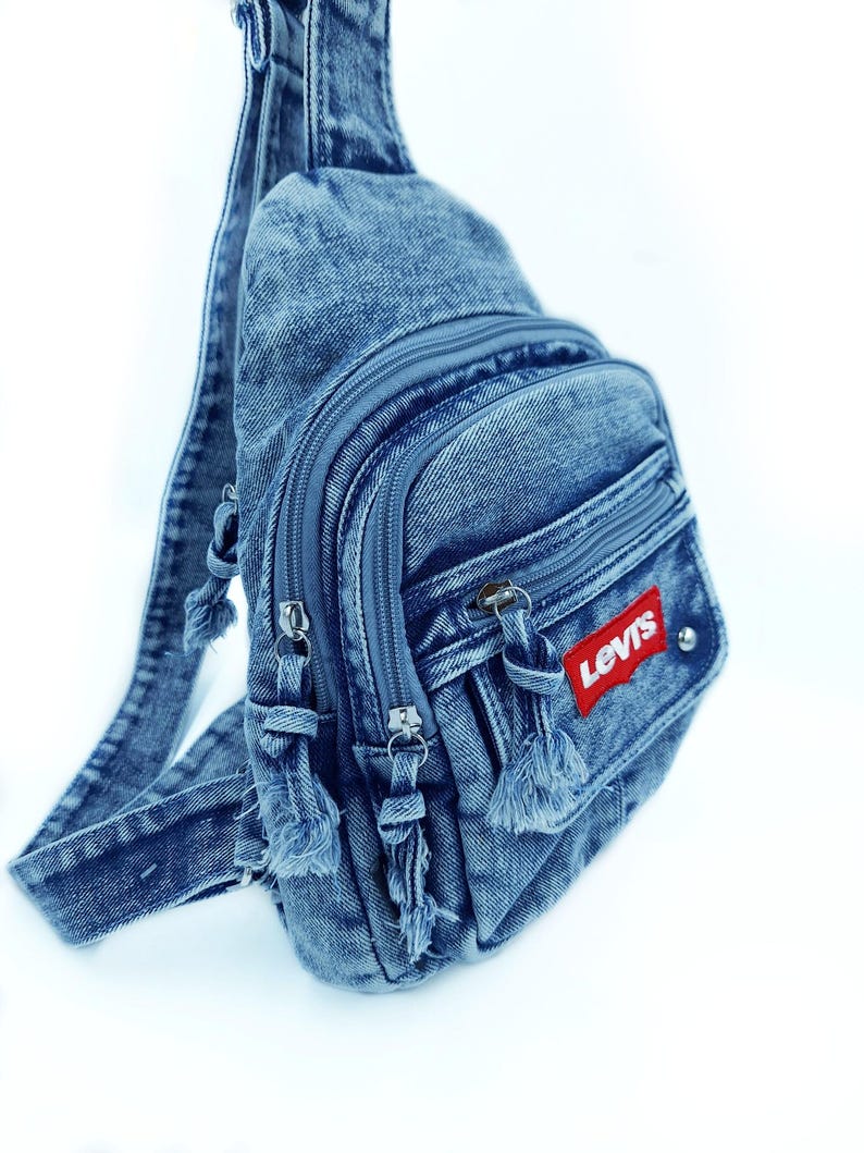 K&ouml;nnte beinhalten: Eine blaue Jeans-Umh&auml;ngetasche mit mehreren Rei&szlig;verschlussf&auml;chern und einem Schultergurt. Die Tasche hat einen roten "Levis"-Aufn&auml;her und dekorative Quasten an den Rei&szlig;verschl&uuml;ssen. Aus Denim gefertigt.