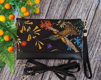 May include: Black clutch bag featuring a vibrant hummingbird and floral pattern. It includes a black wrist strap and a detachable shoulder strap. The base of the bag is black. The background is a wooden surface with orange fruit and green leaves.
