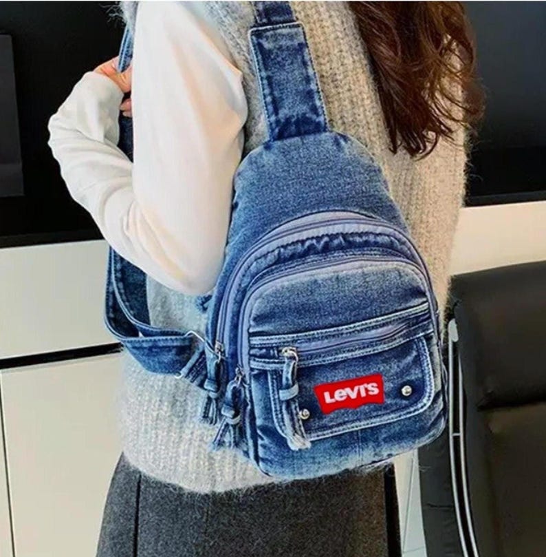 K&ouml;nnte beinhalten: Blauer Jeans-Umh&auml;ngebeutel mit mehreren Rei&szlig;verschlussf&auml;chern und einer Vordertasche. Die Tasche hat ein rotes rechteckiges Levi's-Logo-Patch. Sie hat einen einzelnen verstellbaren Riemen und wird quer &uuml;ber dem K&ouml;rper getragen.