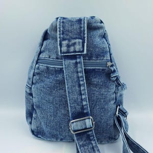 K&ouml;nnte beinhalten: Ein blauer Jeans-Rucksack mit einem einzelnen Schultergurt und einem Rei&szlig;verschluss. Der Rucksack hat eine abgerundete Form und eine kleine obere Klappe. Der Gurt hat eine Metallschnalle zur Einstellung. Der Jeansstoff hat einen verblichenen Look.