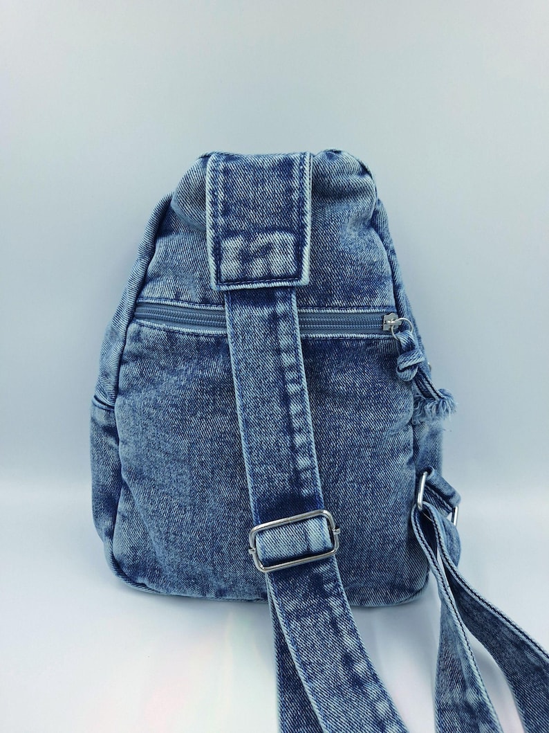 K&ouml;nnte beinhalten: Ein blauer Jeans-Rucksack mit einem einzelnen Schultergurt und einem Rei&szlig;verschluss. Der Rucksack hat eine abgerundete Form und eine kleine obere Klappe. Der Gurt hat eine Metallschnalle zur Einstellung. Der Jeansstoff hat einen verblichenen Look.