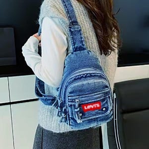 K&ouml;nnte beinhalten: Eine blaue Jeans-Umh&auml;ngetasche mit einem roten "LEVIS"-Patch. Die Tasche hat eine Vordertasche, ein Hauptfach mit Rei&szlig;verschluss und verstellbare Tr&auml;ger. Die Tasche ist aus blauem Jeansstoff.