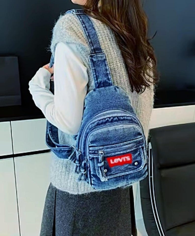 K&ouml;nnte beinhalten: Eine blaue Jeans-Umh&auml;ngetasche mit einem roten "LEVIS"-Patch. Die Tasche hat eine Vordertasche, ein Hauptfach mit Rei&szlig;verschluss und verstellbare Tr&auml;ger. Die Tasche ist aus blauem Jeansstoff.