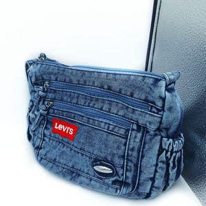 May include: A blue denim crossbody bag featuring multiple zipped compartments, a red Levi's brand patch on the front, and a small silver-toned oval accent. The bag has gathered side pockets and a top zip closure with matching blue fabric pulls.