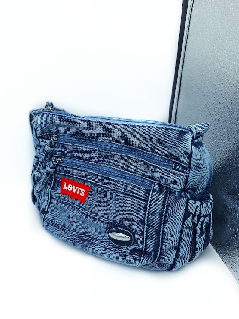 May include: A blue denim crossbody bag featuring multiple zipped compartments, a red Levi's brand patch on the front, and a small silver-toned oval accent. The bag has gathered side pockets and a top zip closure with matching blue fabric pulls.