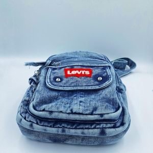 K&ouml;nnte beinhalten: Eine blaue Jeans-Umh&auml;ngetasche mit einer Vordertasche und einem Rei&szlig;verschluss. Die Tasche hat einen roten Levi's-Logoaufn&auml;her. Sie besteht aus Jeansstoff und hat einen Schulterriemen.