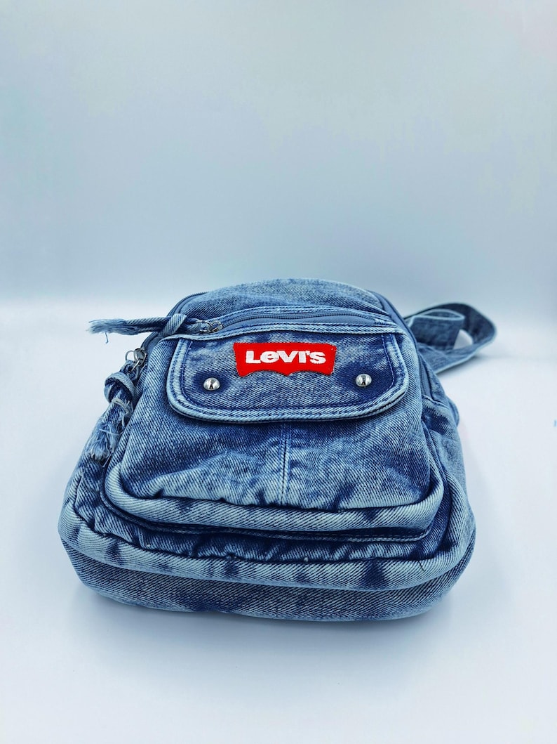 K&ouml;nnte beinhalten: Eine blaue Jeans-Umh&auml;ngetasche mit einer Vordertasche und einem Rei&szlig;verschluss. Die Tasche hat einen roten Levi's-Logoaufn&auml;her. Sie besteht aus Jeansstoff und hat einen Schulterriemen.