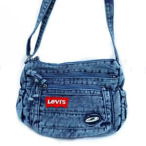 May include: A blue acid-wash denim crossbody bag featuring a red rectangular Levi's brand patch on the front. The bag includes two zippered front compartments, a main zippered opening, side pockets, and an adjustable denim shoulder strap.