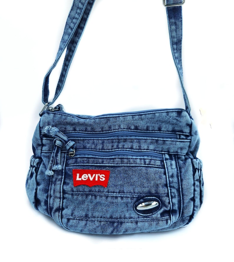 May include: A blue acid-wash denim crossbody bag featuring a red rectangular Levi's brand patch on the front. The bag includes two zippered front compartments, a main zippered opening, side pockets, and an adjustable denim shoulder strap.