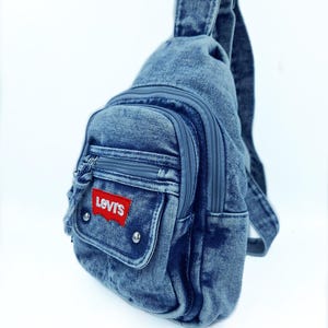 K&ouml;nnte beinhalten: Jeans-Umh&auml;ngetasche mit einer Vordertasche und einem Hauptfach mit Rei&szlig;verschluss. Die Tasche hat einen roten Levi's-Logoaufn&auml;her und silberfarbene Nieten. Sie hat einen einzelnen verstellbaren Riemen und einen oberen Griff.
