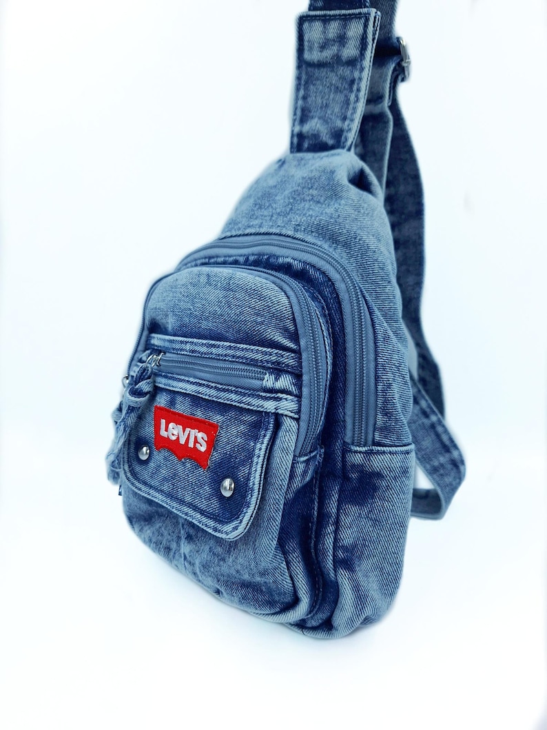 K&ouml;nnte beinhalten: Jeans-Umh&auml;ngetasche mit einer Vordertasche und einem Hauptfach mit Rei&szlig;verschluss. Die Tasche hat einen roten Levi's-Logoaufn&auml;her und silberfarbene Nieten. Sie hat einen einzelnen verstellbaren Riemen und einen oberen Griff.