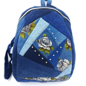 Può includere: Zaino in denim blu con design patchwork. La parte anteriore presenta un motivo floreale con rose bianche e strass. Lo zaino ha un manico superiore, spallacci regolabili e chiusura con cerniera.