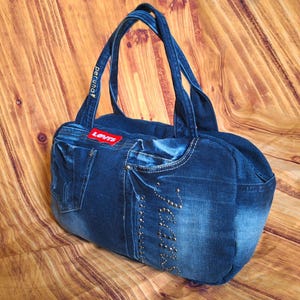 May include: A blue denim tote bag with two handles. The bag features a red "LEVIS" patch and decorative stitching. The bag is made from recycled denim and has a pocket on the side.