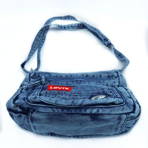 May include: A blue denim crossbody bag featuring a rectangular shape, a long adjustable shoulder strap, and multiple zipped compartments. A red rectangular patch with the white text Levi's is stitched onto the front pocket, accompanied by a small silver-toned metal accent.