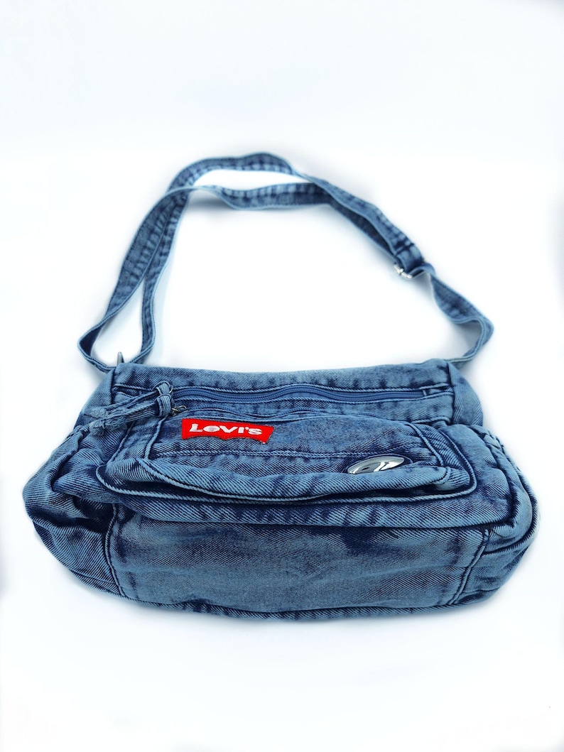 May include: A blue denim crossbody bag featuring a rectangular shape, a long adjustable shoulder strap, and multiple zipped compartments. A red rectangular patch with the white text Levi's is stitched onto the front pocket, accompanied by a small silver-toned metal accent.