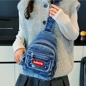 K&ouml;nnte beinhalten: Blaue Jeans-Umh&auml;ngetasche mit Fronttasche und rotem Levi's-Logo. Die Tasche hat einen Rei&szlig;verschluss und ein kleines Quastendetail. Die Tasche wird &uuml;ber der Schulter getragen.