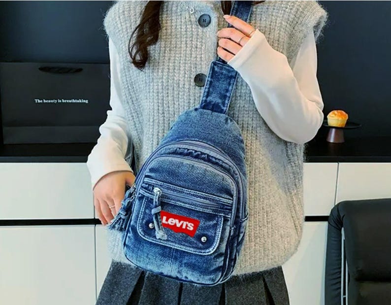 K&ouml;nnte beinhalten: Blaue Jeans-Umh&auml;ngetasche mit Fronttasche und rotem Levi's-Logo. Die Tasche hat einen Rei&szlig;verschluss und ein kleines Quastendetail. Die Tasche wird &uuml;ber der Schulter getragen.